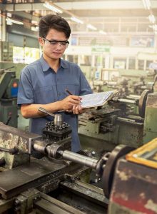 Man with safety glasses inspecting machinery and taking notes on clipboard