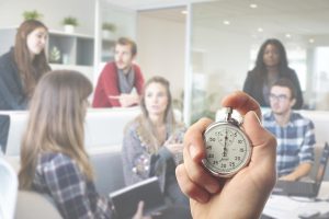 Office scene of a hand holding a stopwatch while coworkers hold a meeting